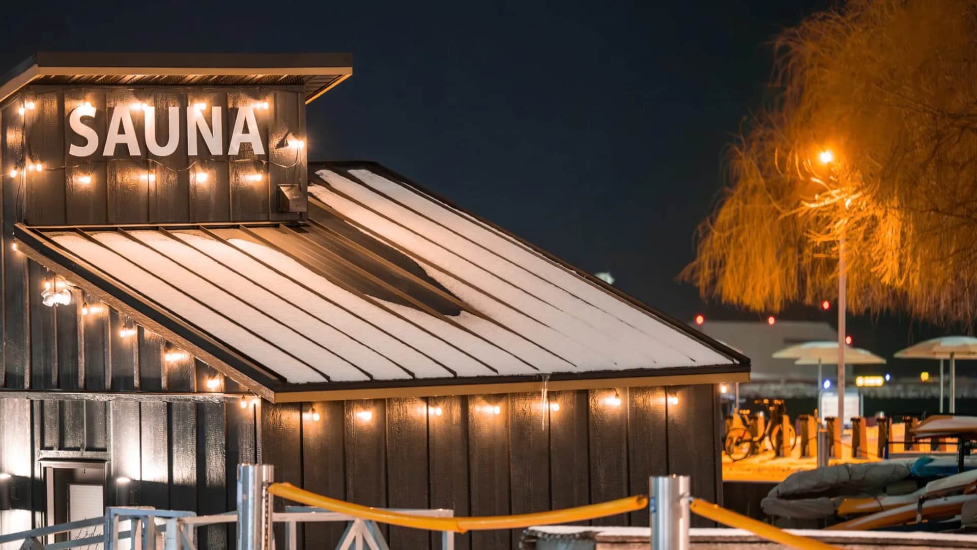 A cozy sauna hut at night, illuminated with string lights, featuring a snow-dusted roof and a warmly lit tree nearby.