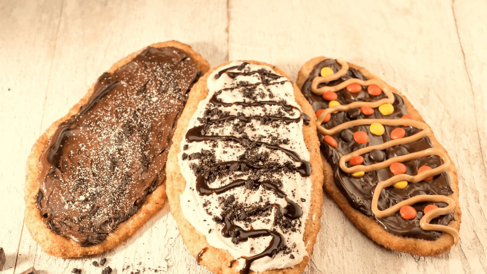 Three different BeaverTails pastries topped with various spreads and toppings, including chocolate and peanut butter, displayed on a rustic wooden surface.