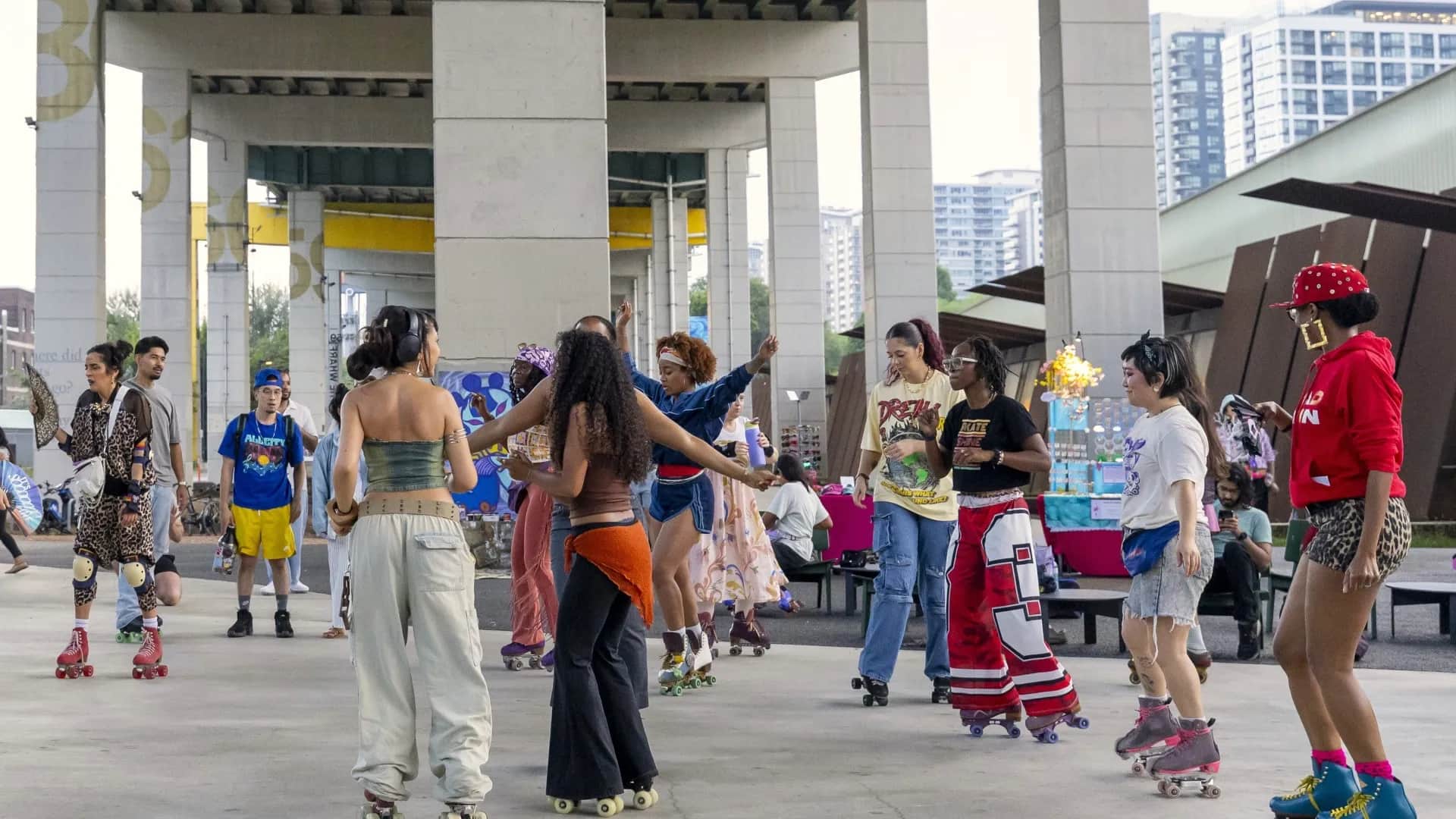 People roller skating and socializing under a highway overpass, with a lively atmosphere and urban backdrop.
