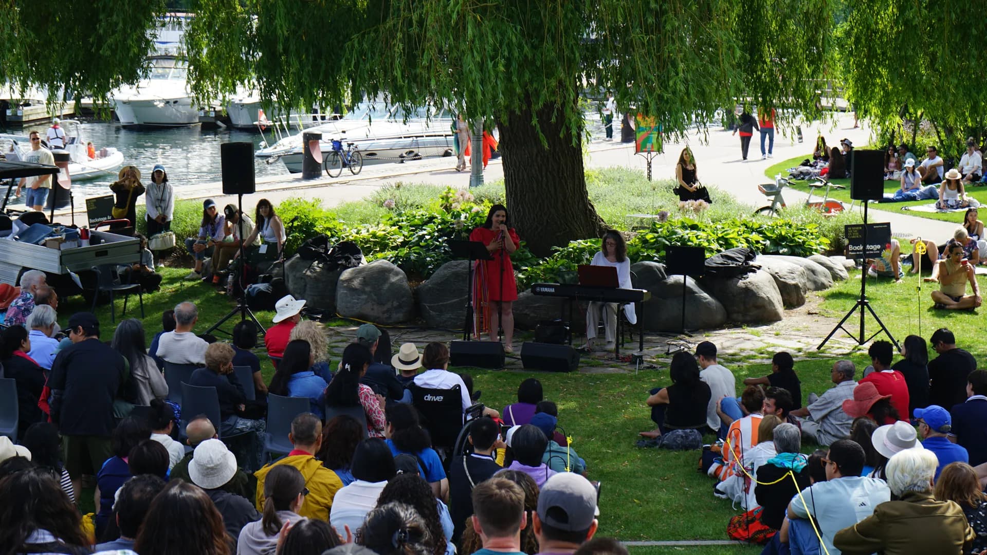 Outdoor concert with a woman singing and another playing keyboard under a large tree. A seated audience watches, with boats docked in the background.