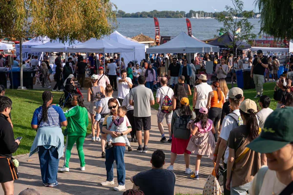 A lively outdoor festival by a waterfront, with diverse crowds exploring vendor booths under white tents on a sunny day.