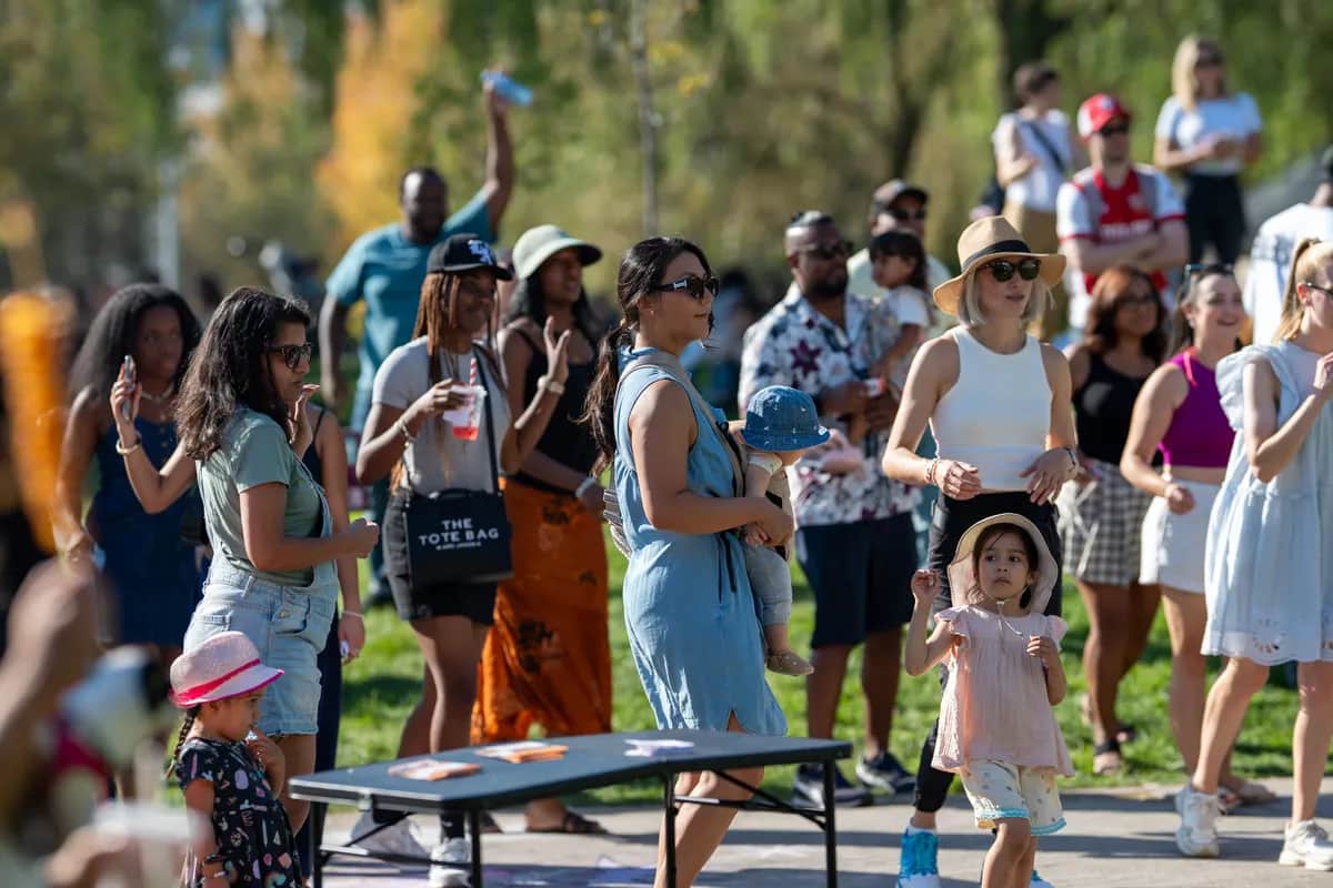 A diverse group of people and children enjoying a sunny outdoor event, some holding drinks and wearing hats, with greenery in the background.