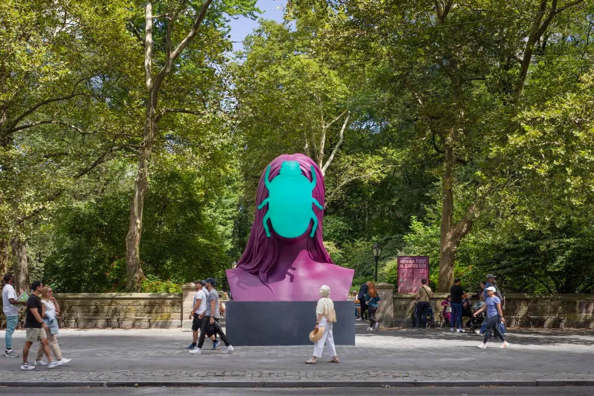 A large, colorful sculpture with a turquoise and purple design stands among trees, while people walk by on a nearby path.