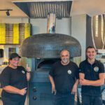 Three employees from Nforno Pizza stand in front of their pizza oven smiling